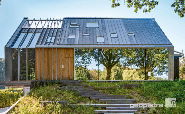 Modern house with large reflective windows, dark sloped roof, and wooden cladding, set in a green environment.